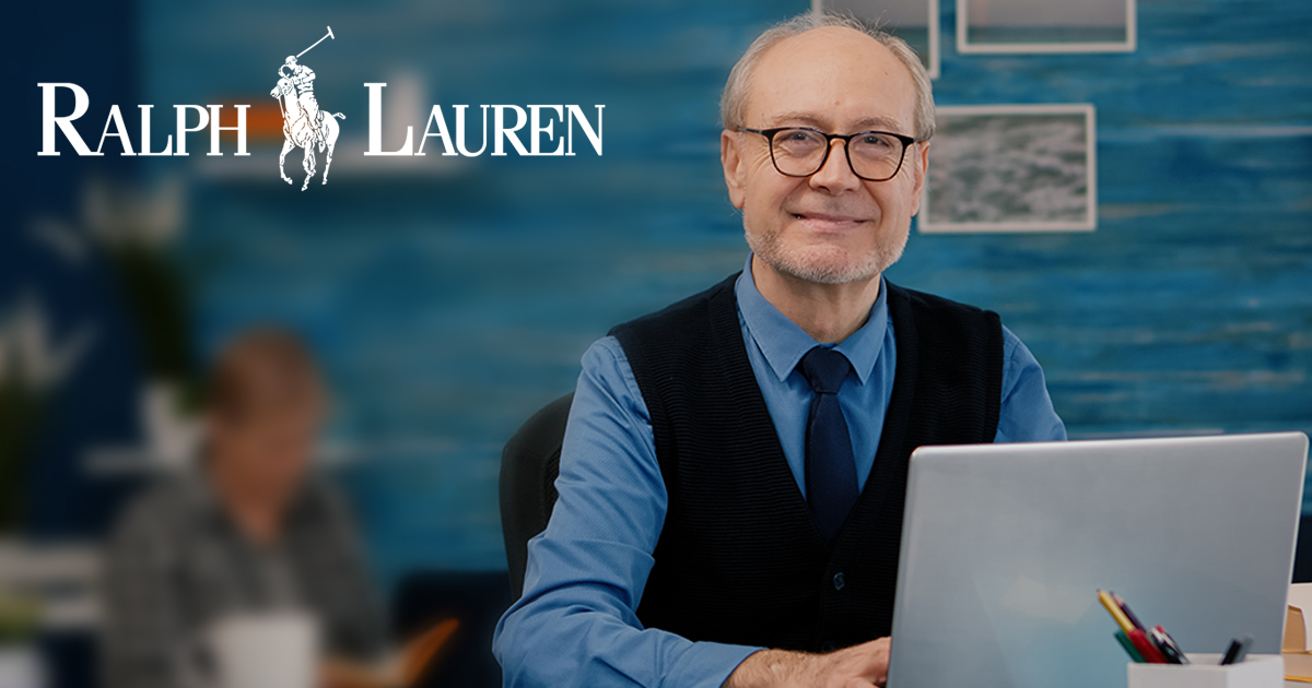 Retired manager sitting at desk in front of camera smiling after typing at laptop working from home while senior wife reading a book in background. Man prepared for online meeting using moden gadgets