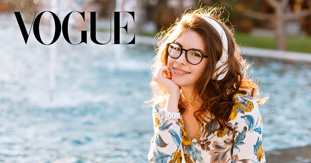 Dreamy girl in white wristwatch and glasses sitting on beautiful fountain propping face by hand. Romantic brown-haired girl listening favorite song in big earphones and looking to camera with smile.